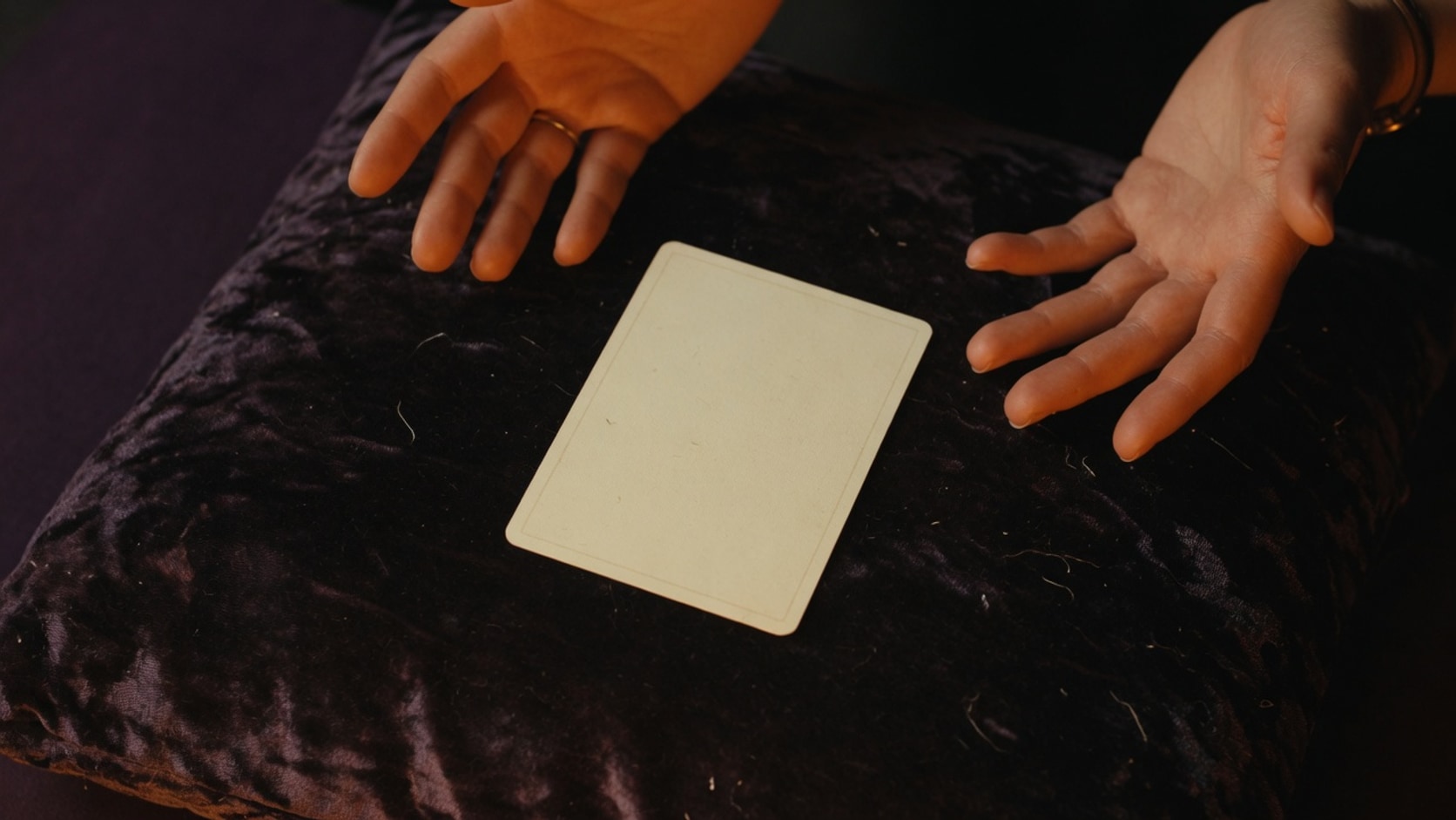AI editorial photograph of a single blank playing card on a velvet cushion between two open hands, left and right