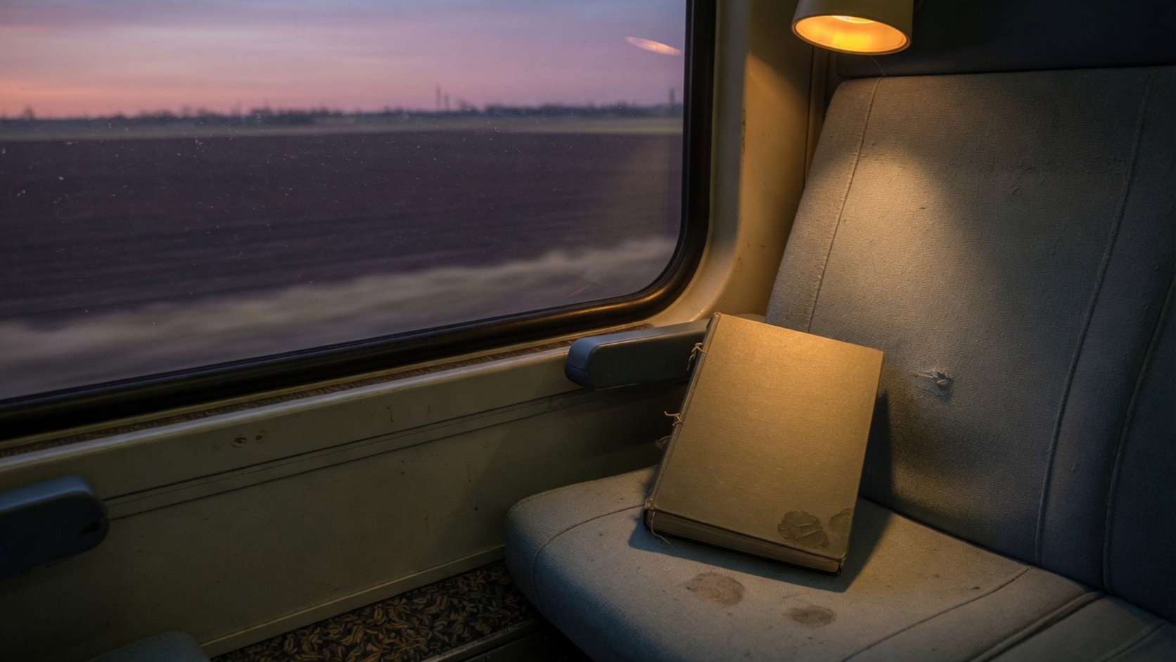 Documentary photograph of a closed unmarked book on a train seat beside a dusk window